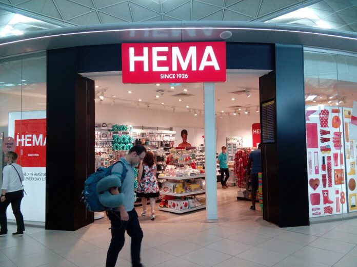 person-wearing-blue-puffer-coat-and-back-pack-walking-past-lit-up-entrance-of-hema-chain-store-with-head-down