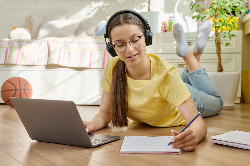 girl-in-headphones-using-laptop-taking-an-online-course-seminar-lying-on-floor-in-her-room-writing-in-notebook