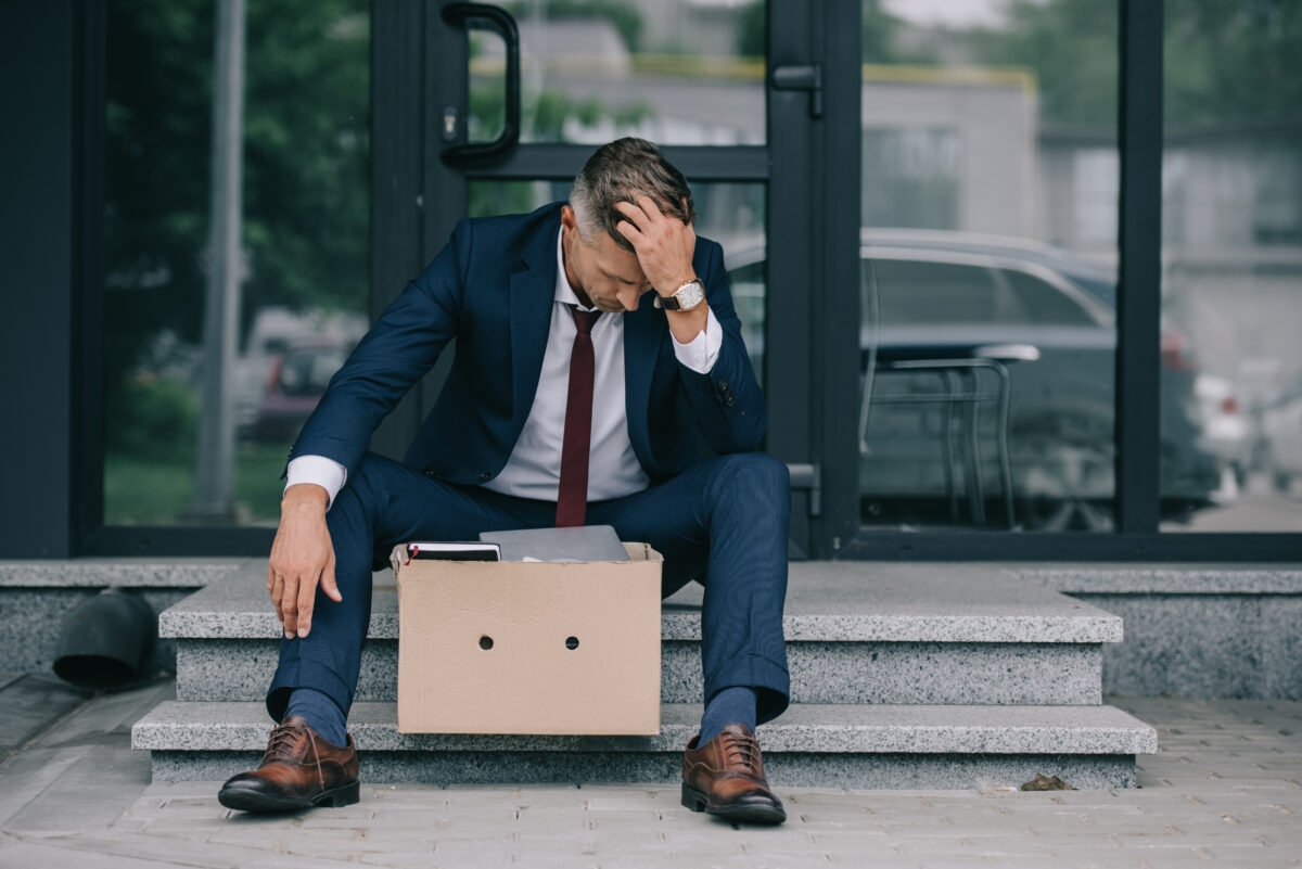 photo of a man sitting on a step with his head in his hand. He has a cartboard box in front of him because he has just been fired from his job.