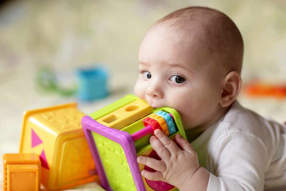 Baby-with-brown-eyes-holding-a-colourful-square-toy-and-putting-it-in-its-mouth