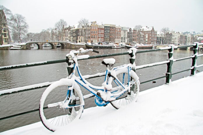 snow-covered-bycicle-leaning-against-canal-rail-with-Amsterdam-houses-in-the-background