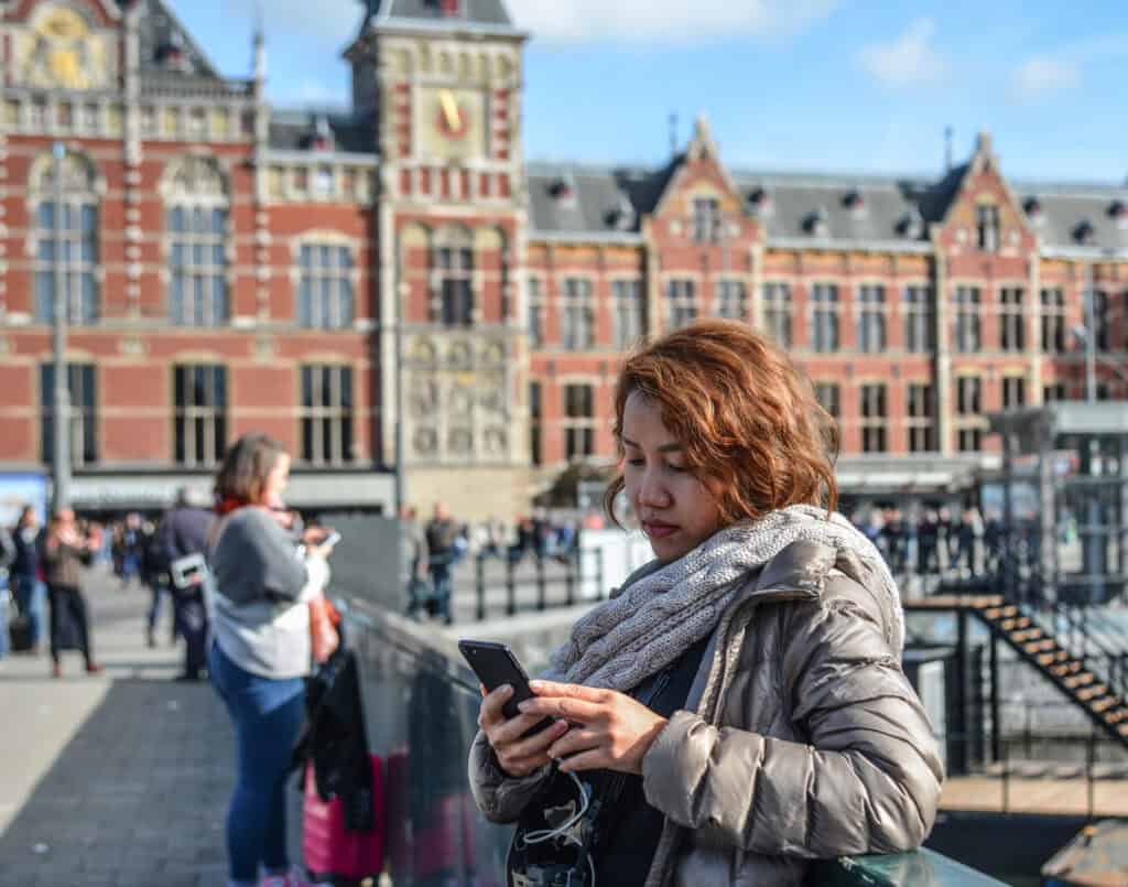 a-woman-stands-outside-centraal-station-in-amsterdam-and-browses-obscure-dating-apps-on-her-phone