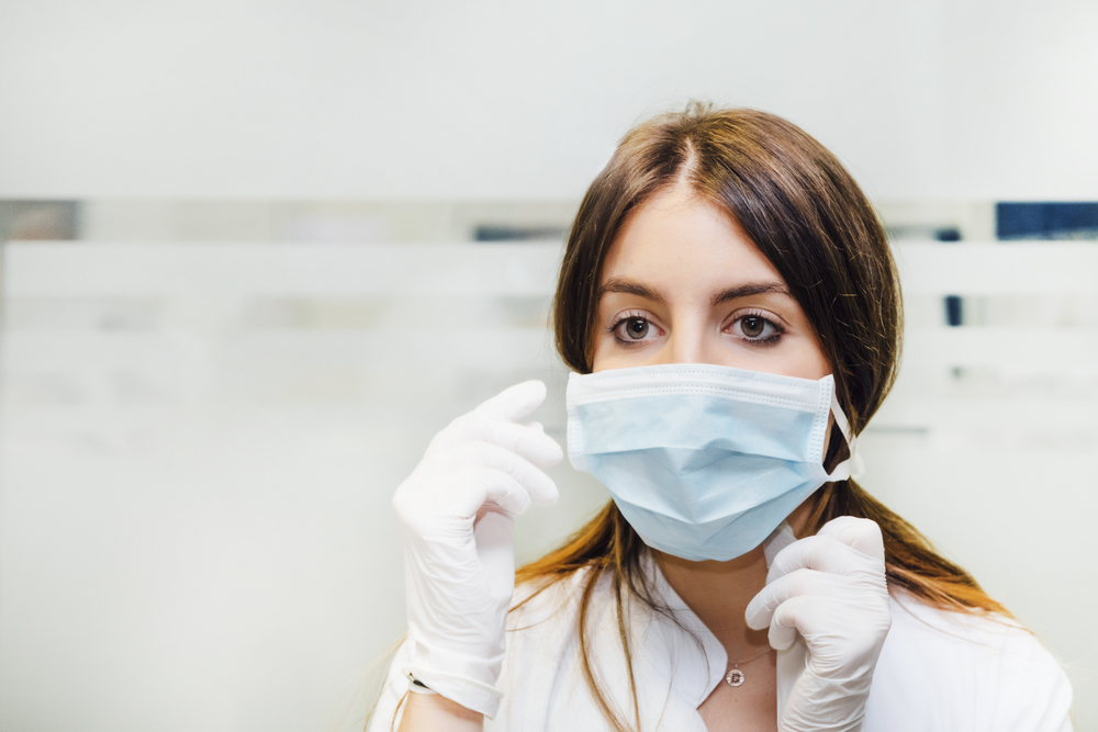 nurse-with-white-gloves-and-brown-air-putting-on-blue-face-mask