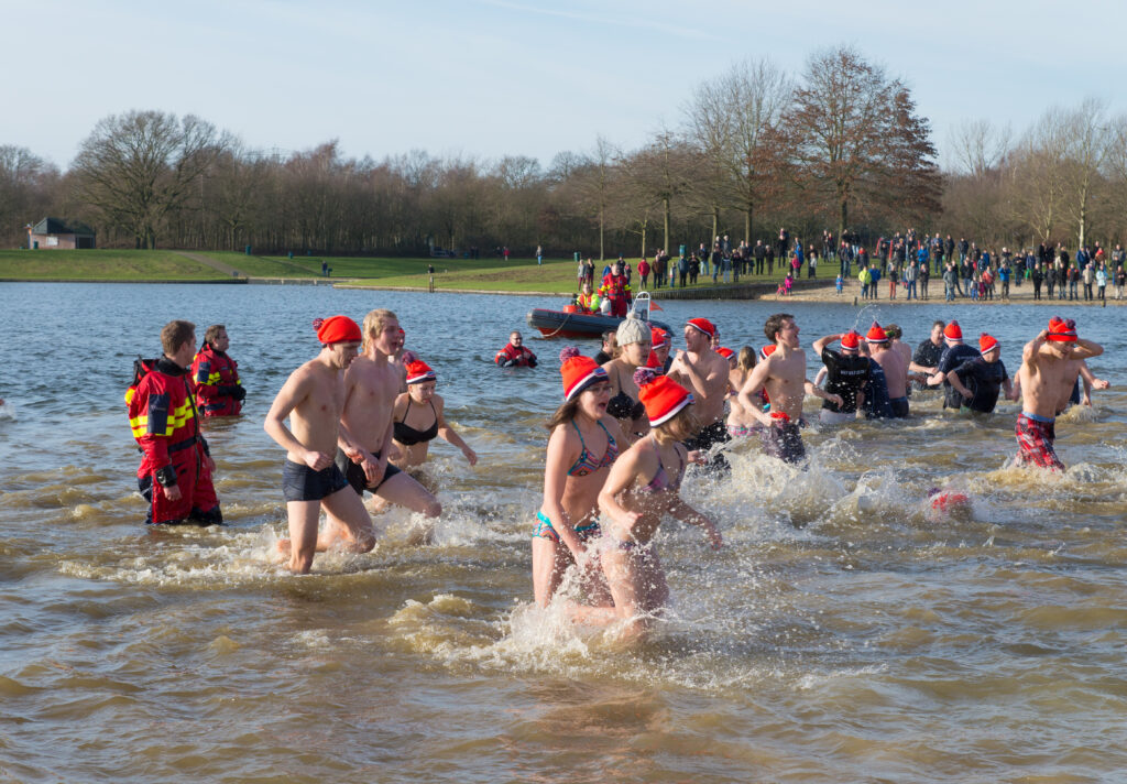 Photo of Dutch people at in water in cold weather. They are wearing an orange hat and swimming trunks, to celebrate the start of the new year. There are lifeguards standing to their left to watch them