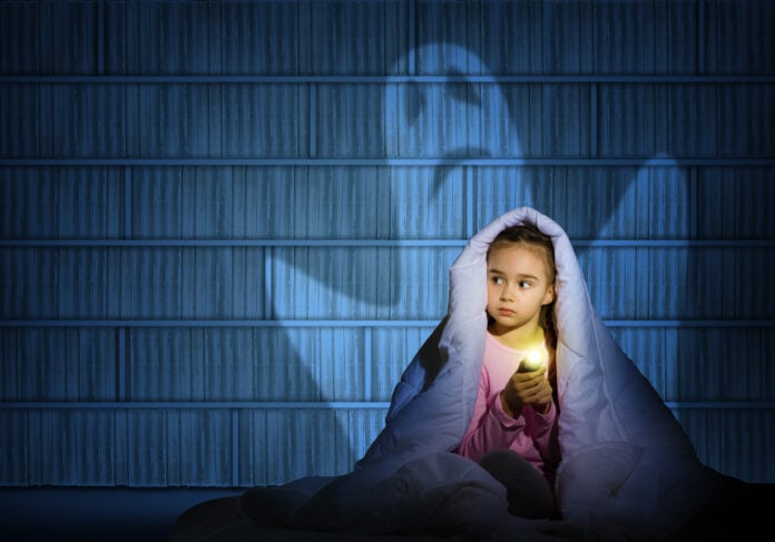 Young-girl-under-covers-holding-a-flashlight-looking-scared-with-ghost-behind-her