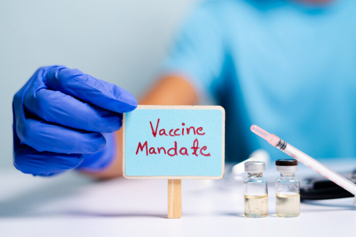hand-of-nurse-with-blue-glove-holding-sign-with-vaccine-mandate-written-on-it-next-to-syringes