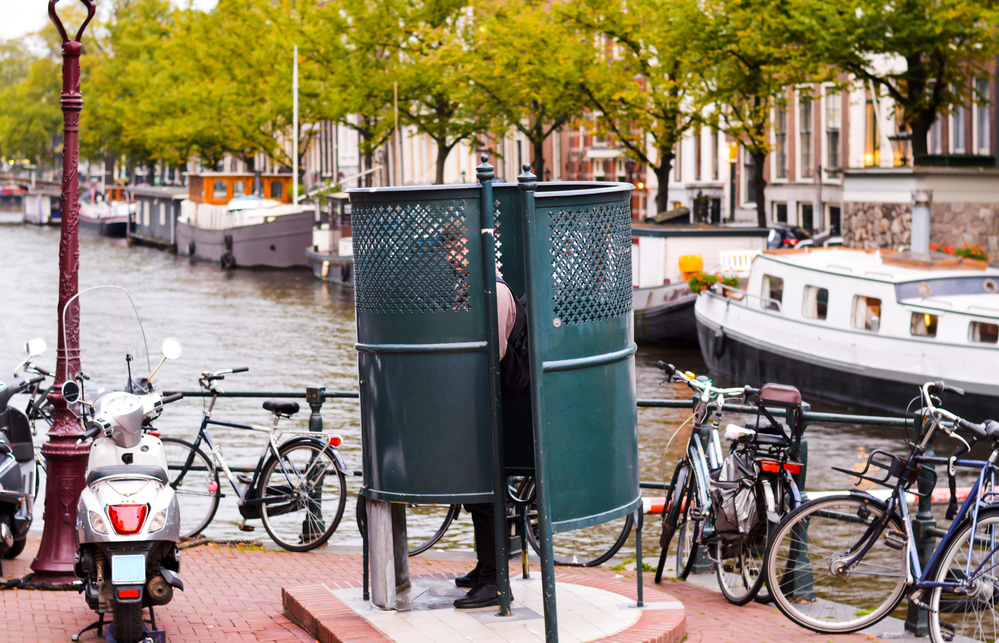 picture-of-a-man-in-public-urinal-in-Amsterdam-surrounded-by-bikes-scooter-boats-background-of-canal-houses-trees