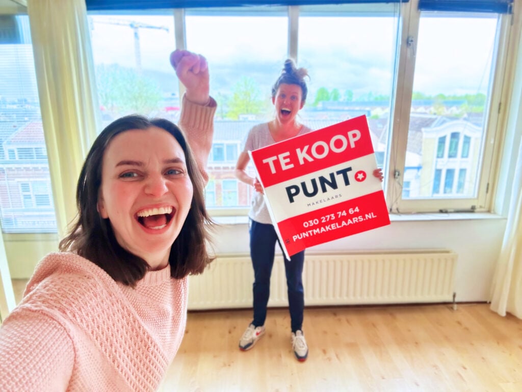 author-and-girlfriend-holding-te-koop-sign-in-new-apartment-with-fists-raised-in-air