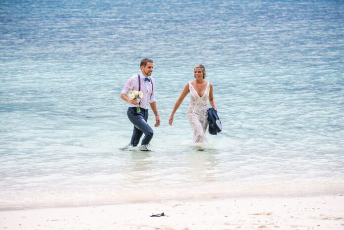 couple-in-wedding-attire-standing-knee-deep-in-tropical-water-walking-towards-beach