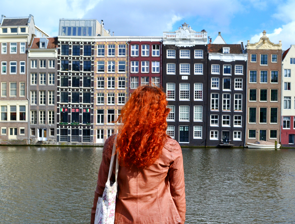 Photo-of-redheaded-woman-standing-at-canal-looking-at-Amsterdam-houses-on-a-sunny-day