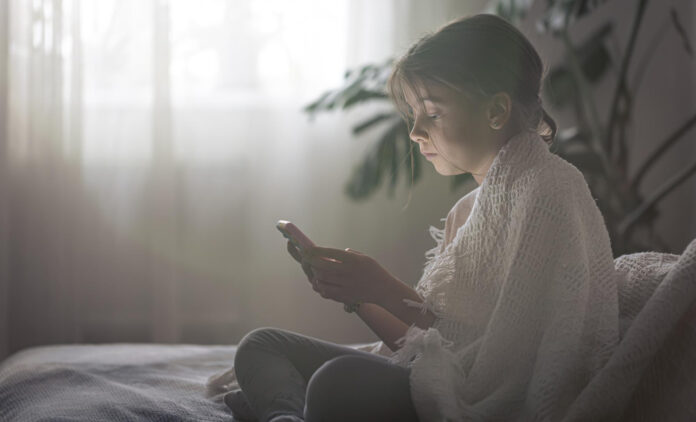 Young-girl-sits-on-her-bed-with-a-blanket-while-using-her-smartphone