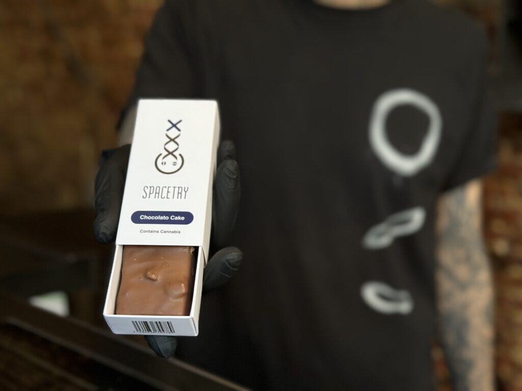 closeup-of-chocolate-cake-edible-in-white-box-being-held-by-budtender-in-black-shirt-with-white-circles-in-amsterdam
