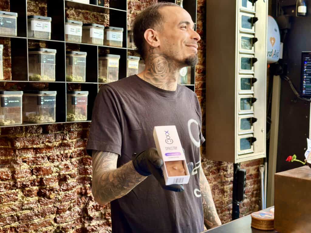 photo-of-budtender-smiling-looking-away-from-camera-and-holding-out-shocolate-cake-edible-with-brick-industrial-background-at-coffeeshop-amsterdam