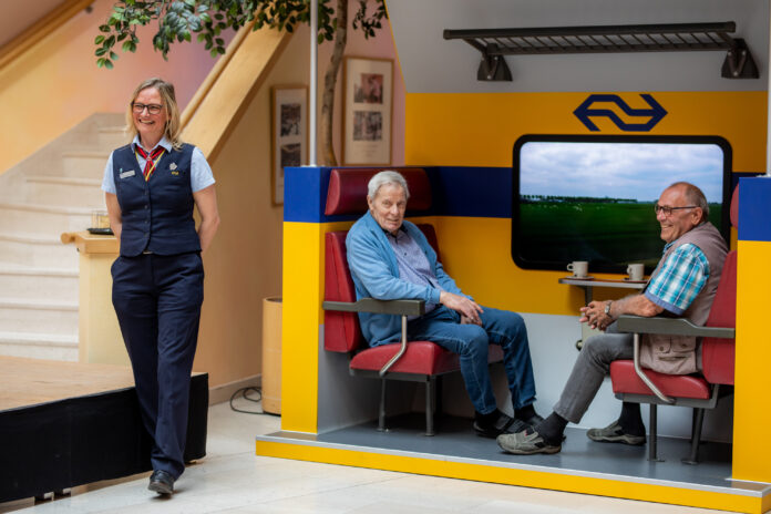 Photo of two elderly men sitting in a small imitation train carriage in the common room of their nursing home. There is a woman dressed in uniform standing next to them.