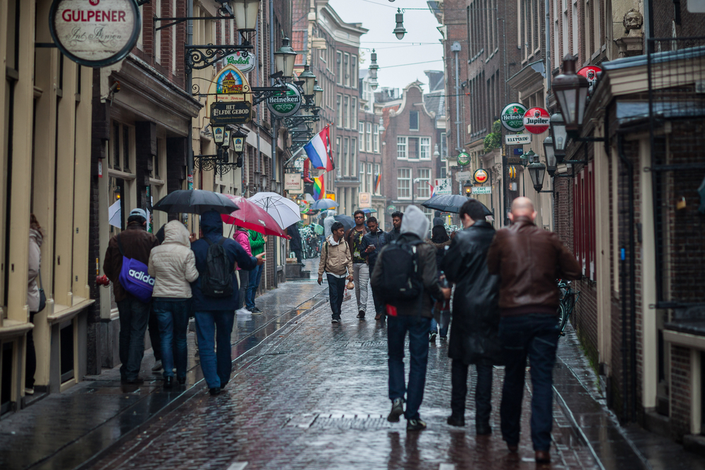 photo-of-many-people-walking-in-rain-in-amsterdam-wet-narrow-street-during-winter-umbrellas-dutch-flag