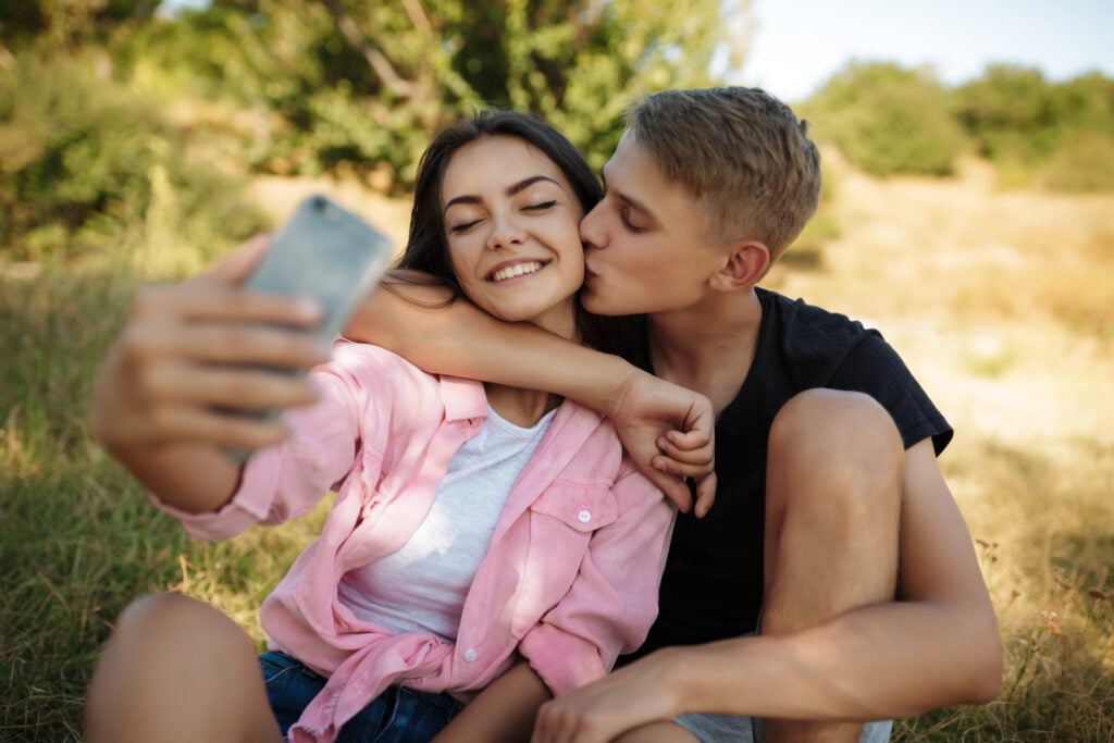 Portrait of young smiling couple sitting on lawn in park and making selfie. Beautiful couple taking photos on cellphone frontal camera in the Netherlands.