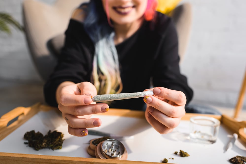 woman-with-colourful-hair-holding-joint-towards-viewer-over-table-with-grinder-and-cannabis-buds