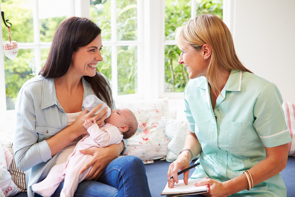 Young-mother-bottle-feeding-her-newborn-smiling-at-her-midwife-with-bright-windows-in-the-background