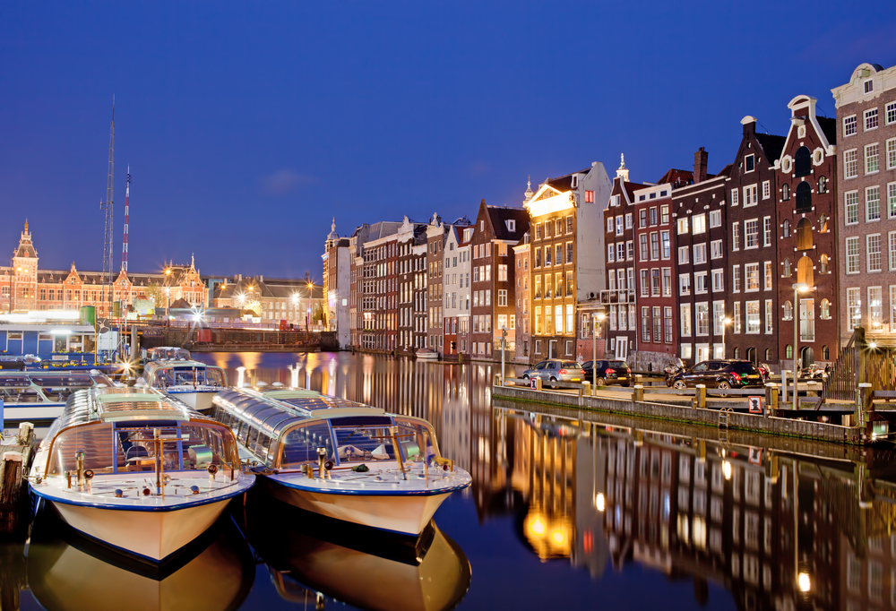 City-of-Amsterdam-in-Netherlands-at-night-historic-apartment-houses-with-reflections-on-water-and-boats-ready-for-canal-tours-and-cruises