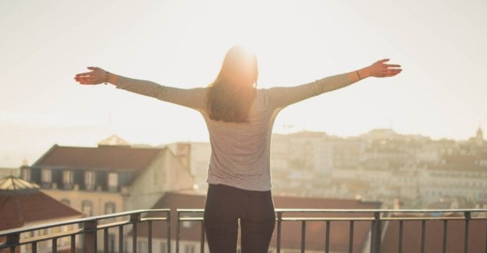 Woman stands on balcony with arms spread open in the sunshine