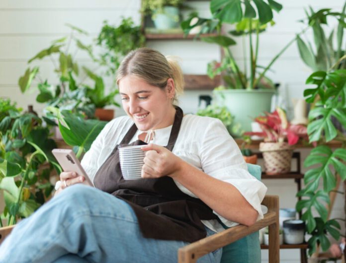 photo-of-small-business-owner-sipping-coffee-looking-at-phone-with-plants-in-background
