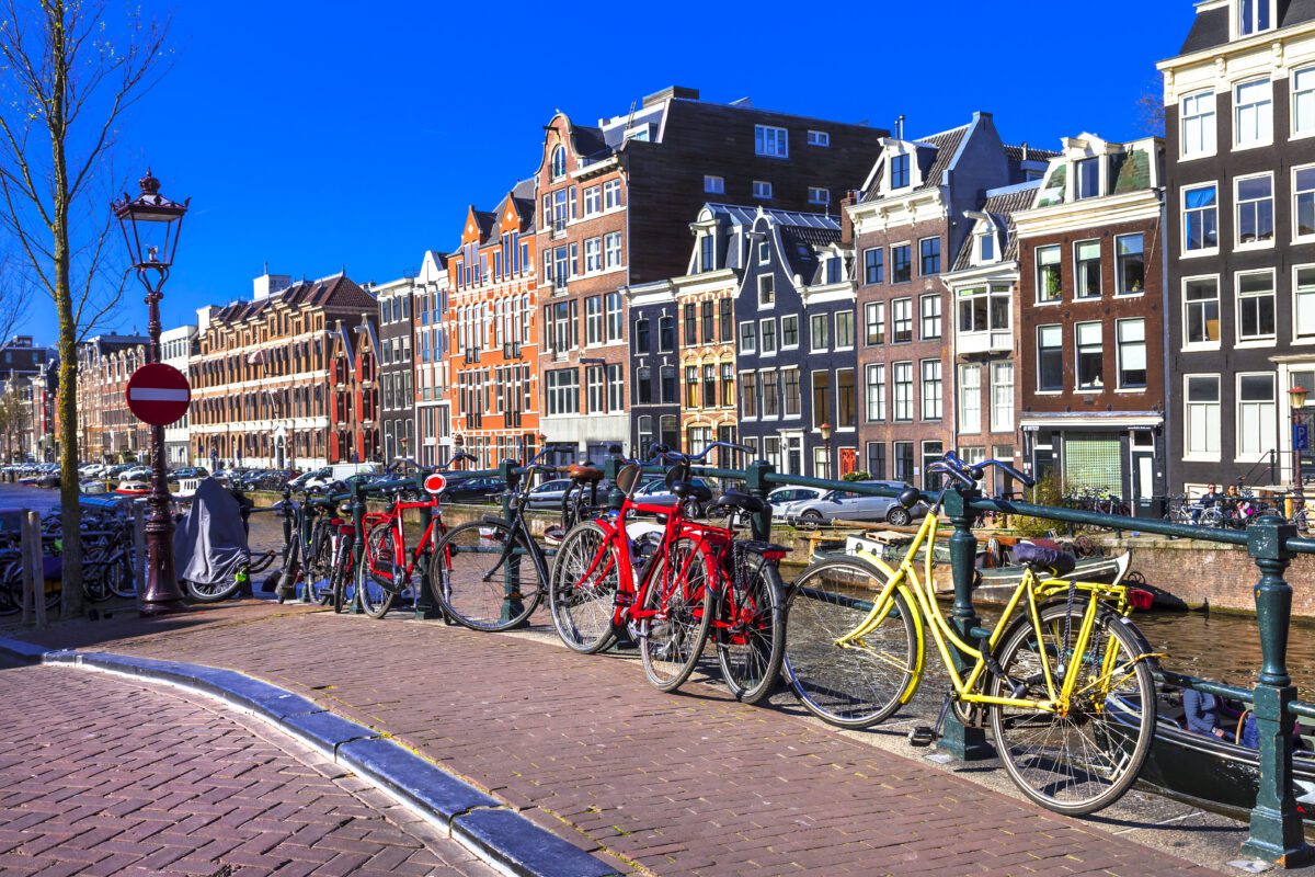 bridge-with-bikes-lined-up-against-it-on-canal-bank-with-blue-sky-and-typical-dutch-houses-in-background-of-amsterdam-in-the-netherlands