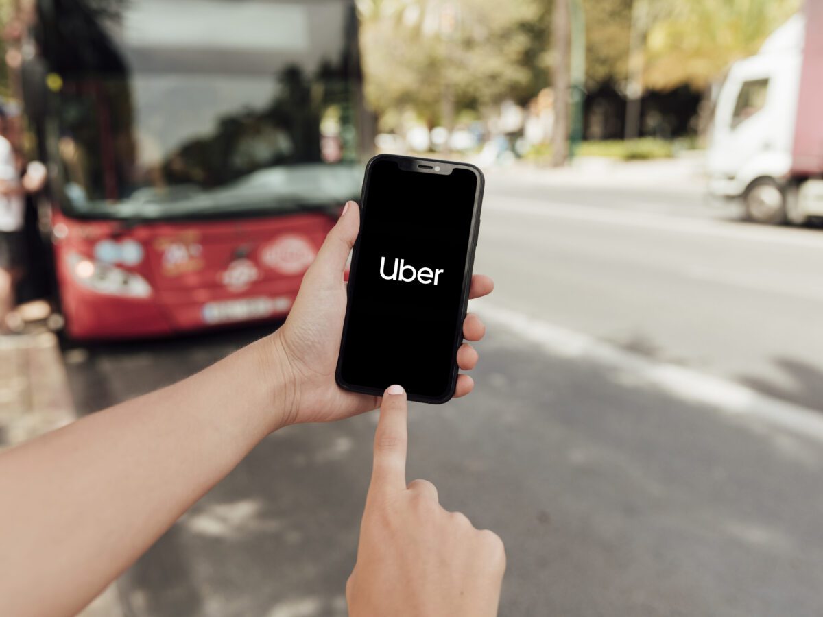 close-up-finger-pointing-to-phone-screen-with-uber-app-open-while-bus-stands-in-background-scaled