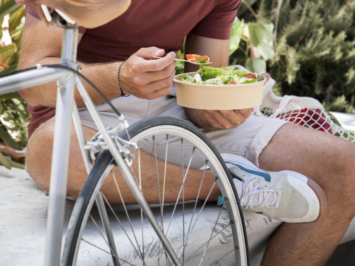 young-man-bent-down-sitting-on-marble-bench-eating-salad-with-bicycle-beside-him