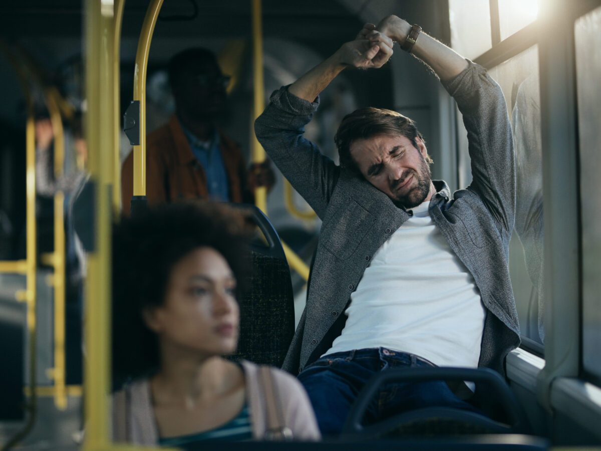 exhausted-adult-man-stretching-while-traveling-by-bus-with-woman-sitting-in-front-of-him-scaled