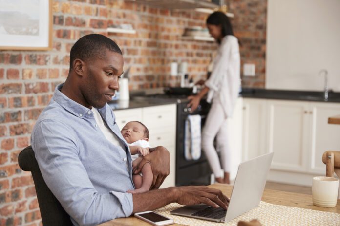 photo-of-man-sitting-on-table-working-from-home-holding-his-child-while-his-wife-cleans-the-house-in-the-background