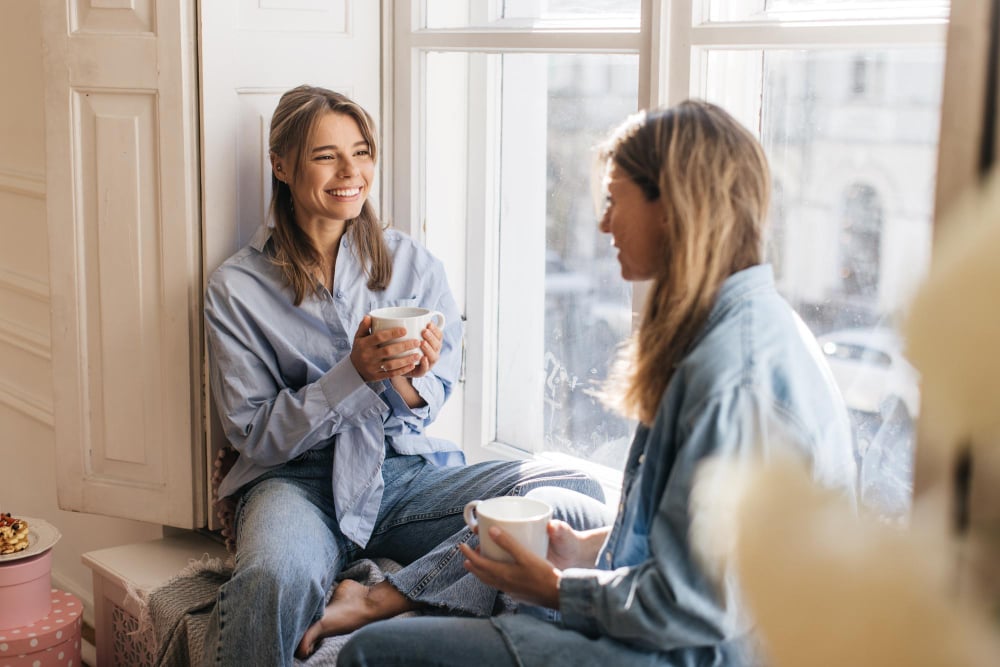 photo-of-two-friends-sitting-by-window-chatting-wtih-coffee
