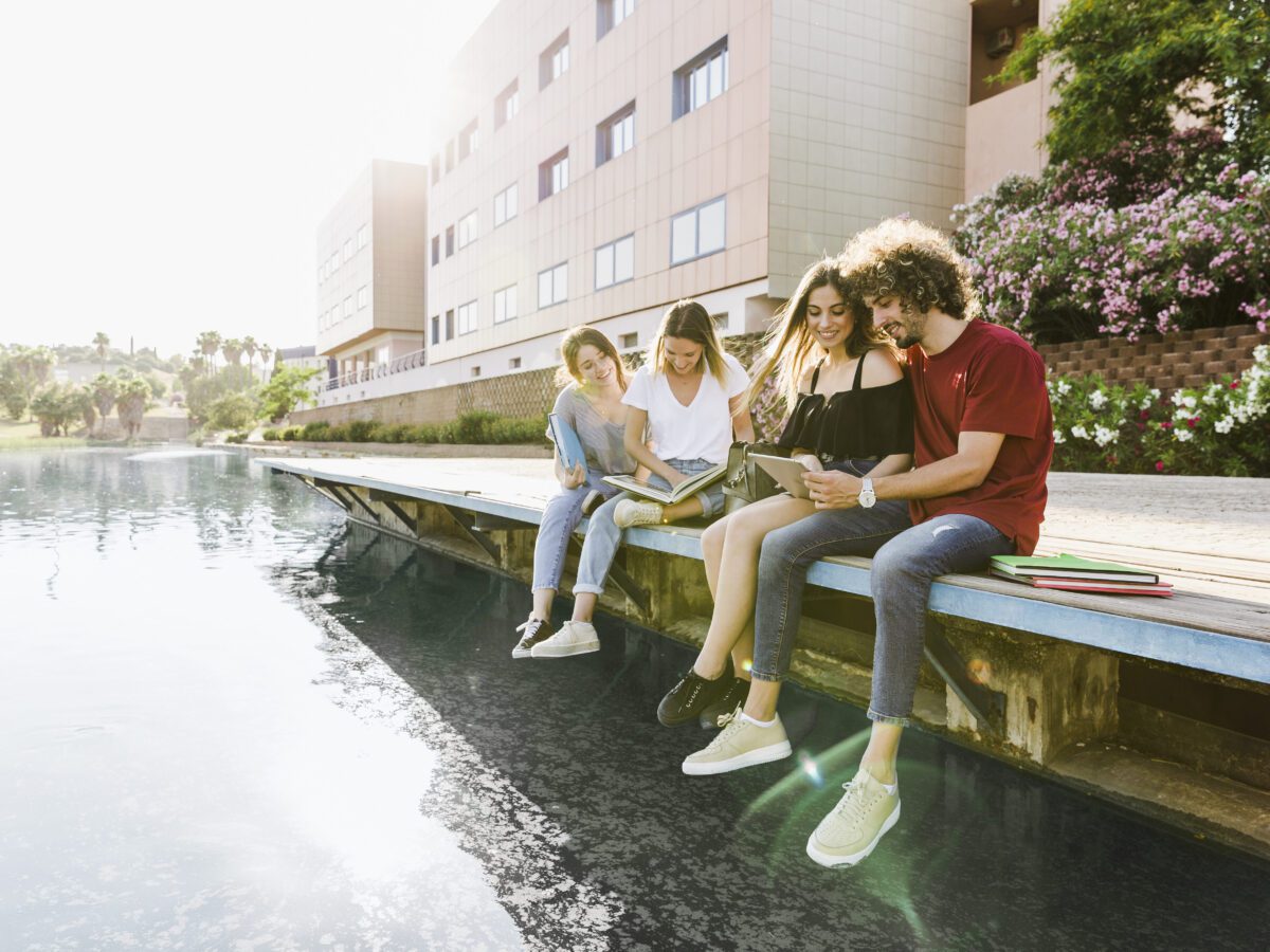 student-friends-smiling-and-studying-near-building-with-legs-dangling-over-lake-scaled