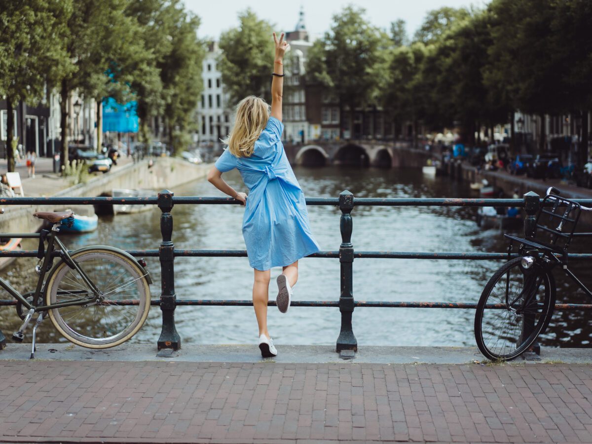 blond-girl-with-blue-dress-and-white-shoes-jumping-up-on-amsterdam-bridge-making-peace-sign-in-the-air