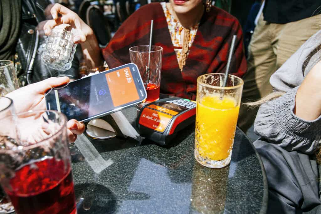 group-of-friends-sitting-at-outside-restaurant-table-with-drinks-while-one-makes-contactless-payment-with-ING-on-phone