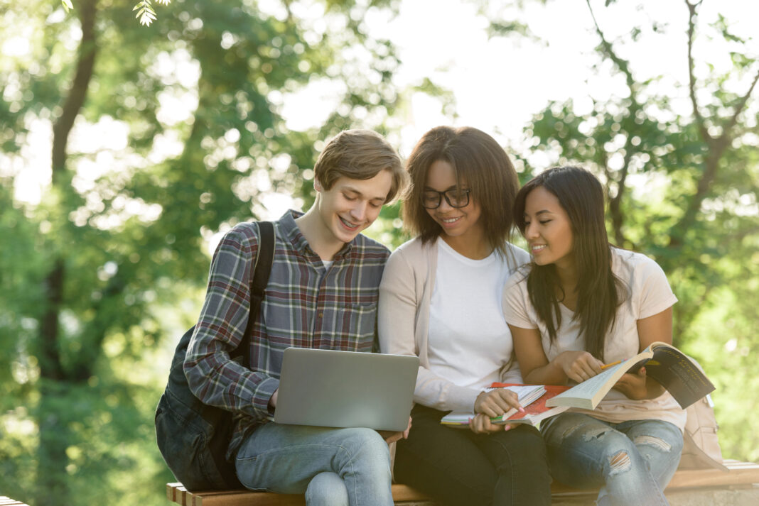Happy-students-sitting-studying-outdoors-while-using-a-laptop
