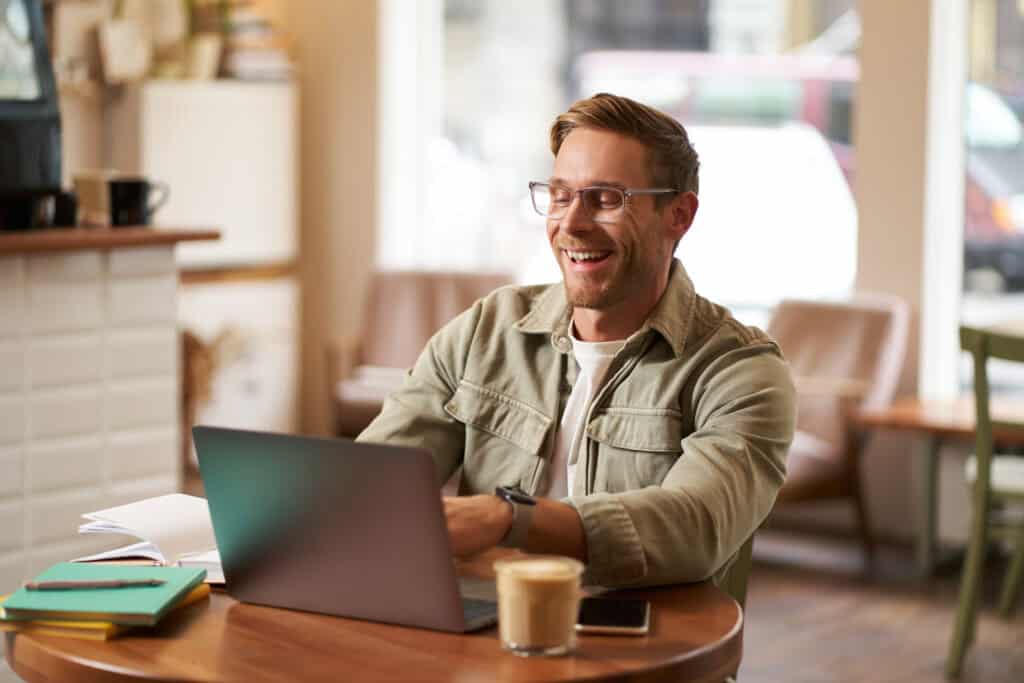 Image of young digital nomad, man in glasses sits in cafe, works from coffee shop, uses laptop in co-working space, wears glasses, drinks his beverage