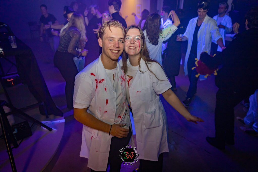 girl-and-boy-dressed-in-bloody-white-lab-coat-costumes-smiling-and-posing-at-party-with-people-dancing-in-background