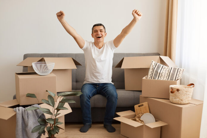 Indoor shot of extremely happy brunette man wearing white T-shirt and jeans sitting on couch surrounded with pile of packages, raised arms, celebrating moving to a new flat or house. Happy that he doesn't have to pay high deposit.