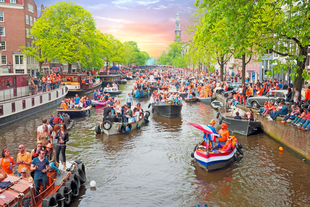 photo-of-boats-crowding-amsterdam-canal-full-of-people-dressed-in-orange-for-kings-day-with-sunset-behind