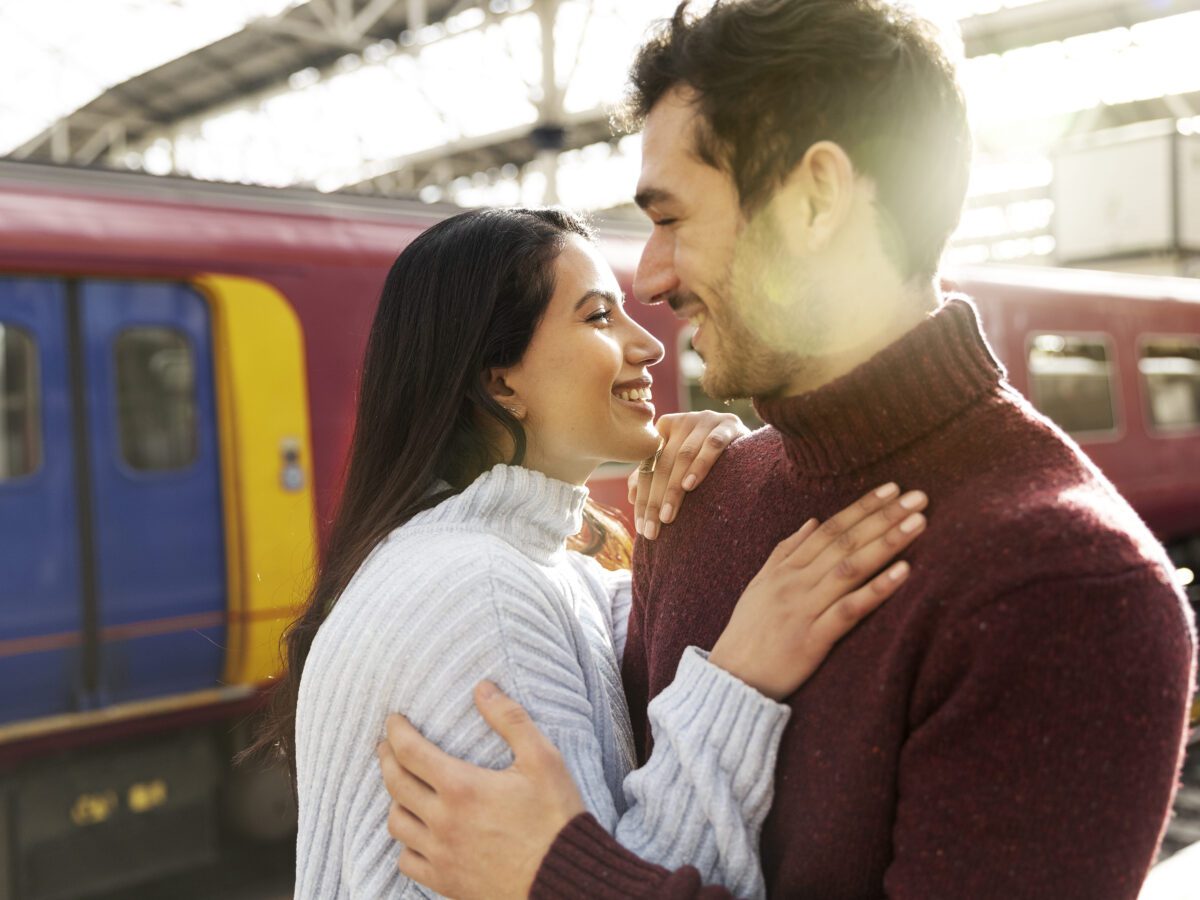 couple-wearing-turtle-necks-holding-each-other-laughing-outside-of-train-platform-with-train-in-background