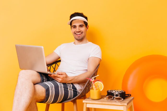 man-wear-cap-and-white-t-shirt-and-striped-shorts-sits-against-orange-background-with-laptop-smiling-with-cocktail-beside-him