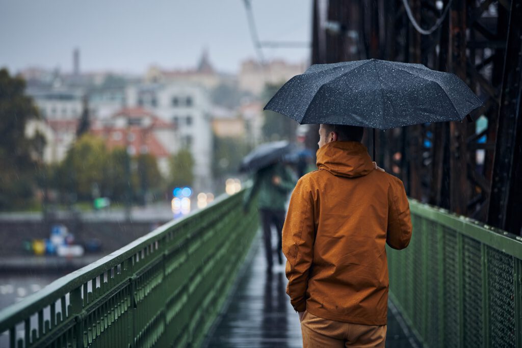 Photo-of-young-man-wearing-orange-raincoat-and-holding-umbrella-while-walking-in-the-rain-on-a-bridge