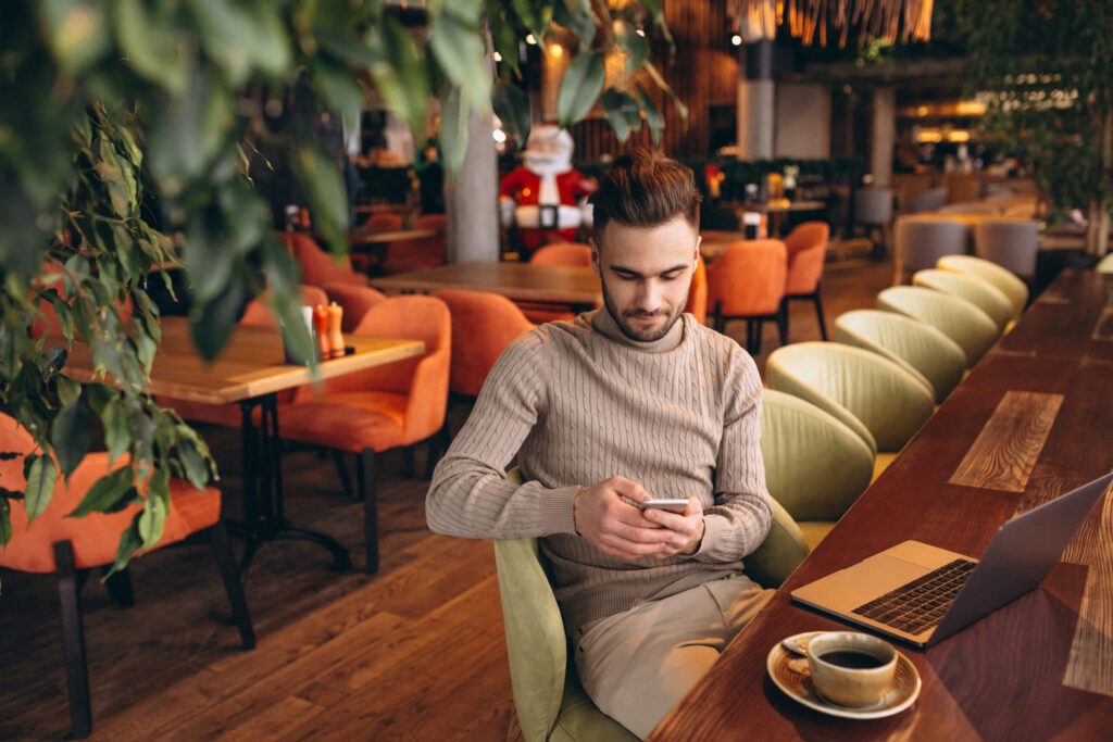 photograph-of-a-Dutch-man-in-a-beige-turtleneck-looking-at-his-phone-using-sim-only-subscription-whilst-his-laptop-and-cup-of-coffee-sit-on-the-cafe-table-next-to-him