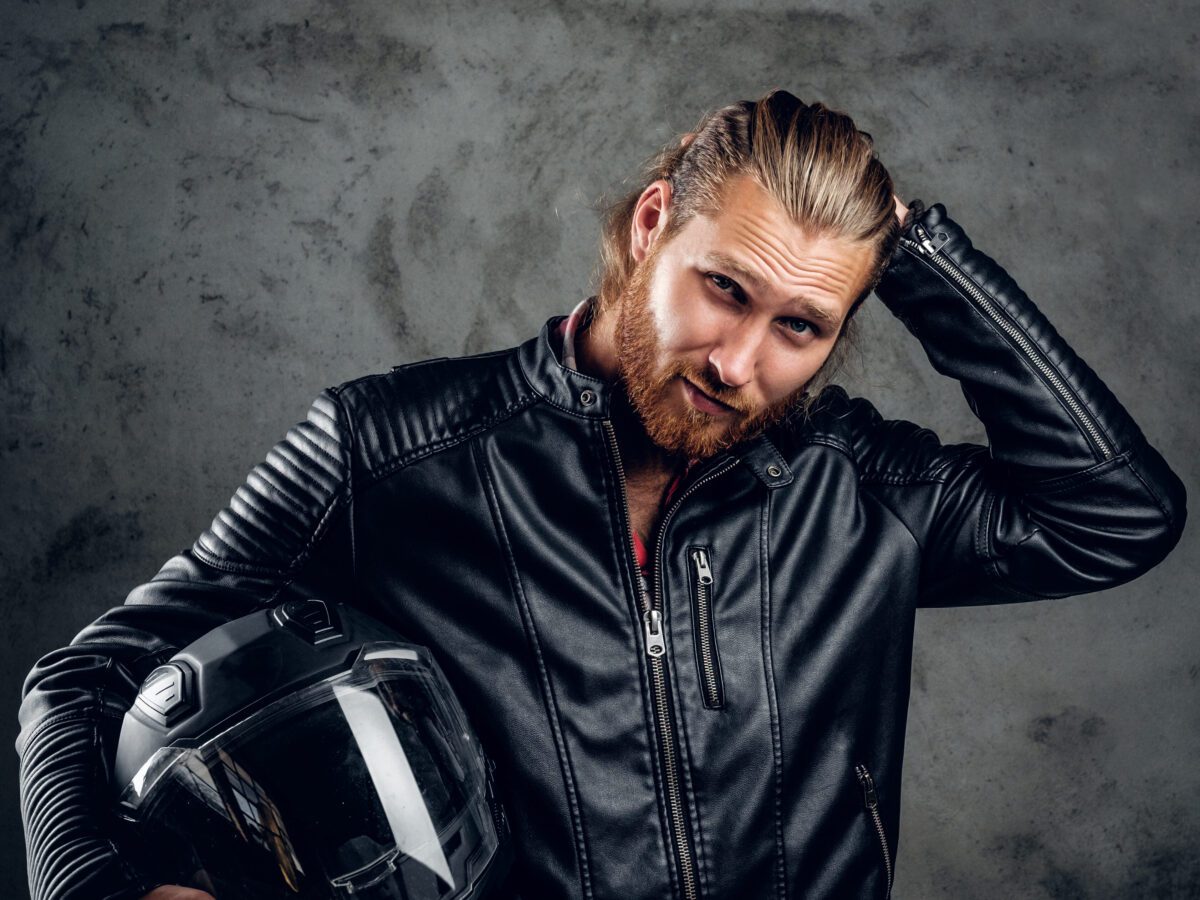 bearded-redhead-male-wearing-leather-jacket-holds-motorcycle-helmet-against-grey-background-scaled