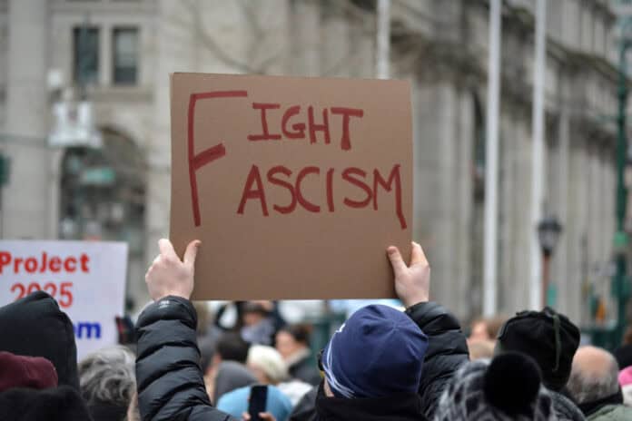 photo-of-man-holding-up-brown-protest-poster-red-writing-fight-fascism-amrs-in-air-crowd-walking-marching