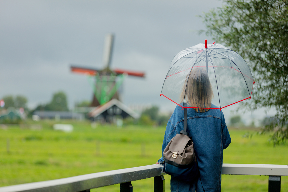 a-picture-of-a-girl-with-an-umbrella-on-a-rainy-day-with-dutch-windmills-wearing-denim-blue-thin-jacket-bag-over-one-shoulder