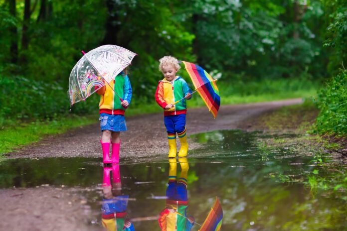 Little boy and girl play in rainy summer park. Children with colorful rainbow umbrella, waterproof boots jump in puddle and mud in the rain. Kids walk in autumn shower. Outdoor fun by any weather
