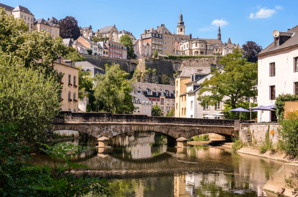 Image of Luxembourg city on a road trip from the Netherlands. We see a bridge over the water, surrounded by trees and houses.