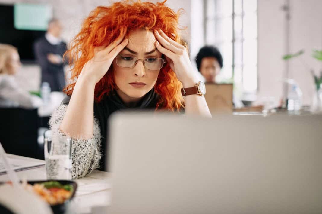 photo-of-a-serious-businesswoman-holding-her-head-reading-problematic-email-on-laptop-while-working-in-the-office-there-are-people-in-the-background
