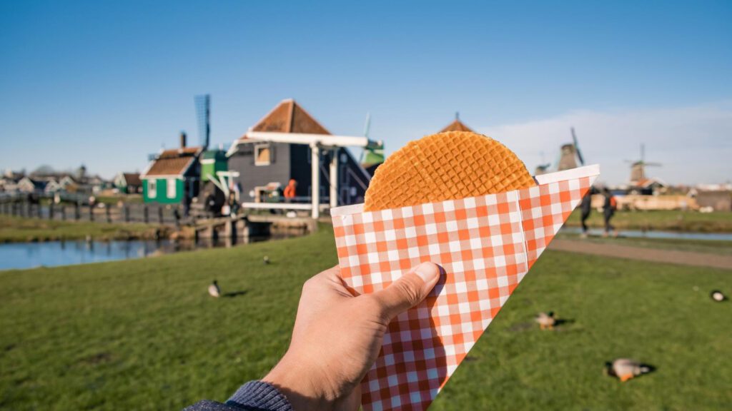 photo-of-person-holding-stroopwafel-in-red-white-checkered-paper-in-front-of-zaanse-schans-windmill-background